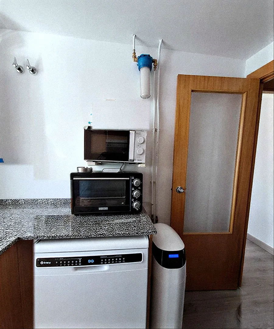 Descalcificador VD compacte instal·lat al costat del rentaplats en una cuina, amb filtre d'entrada a la paret